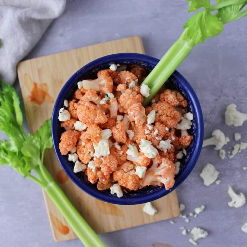 Cauliflower covered in Frank's hot sauce with blue cheese crumbles and a celery stalk sticking out the bowl.