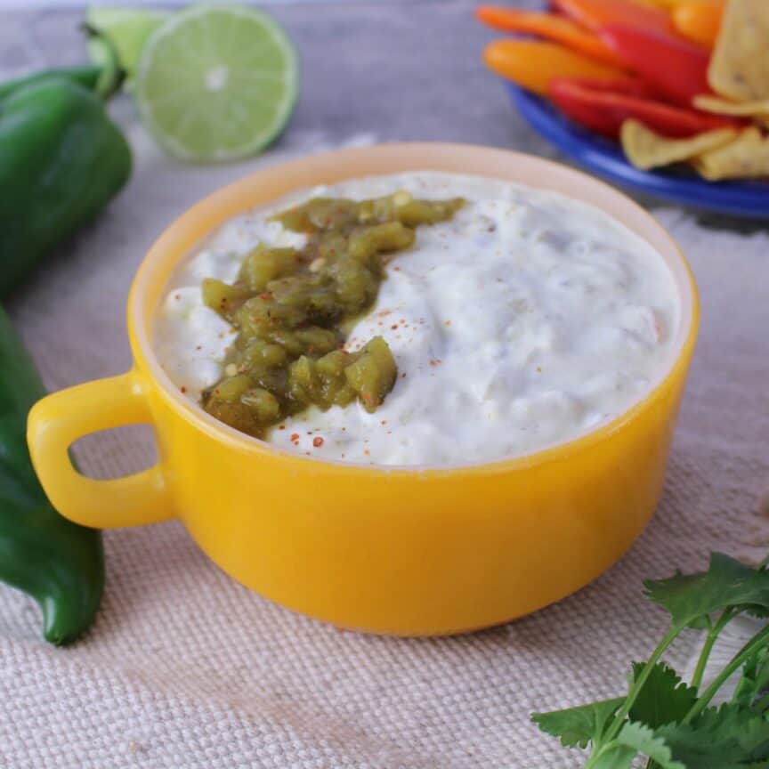 healthy queso in yellow bowl with mini pepper, limes, chilies, and cilantro on the side