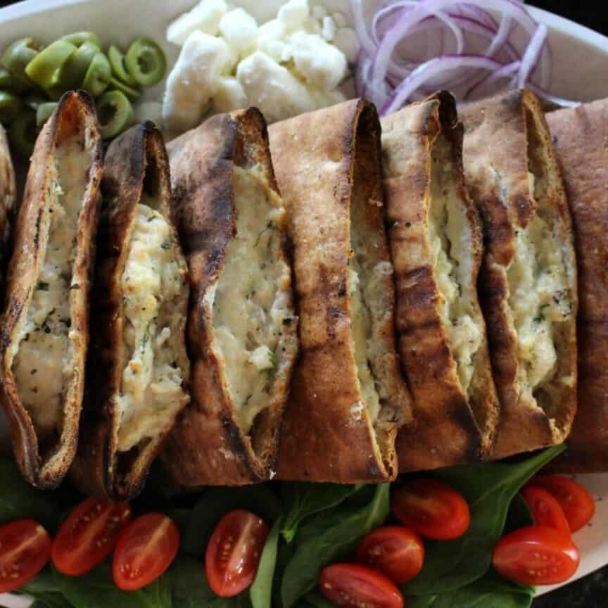 Grilled pita pocket bread that is stuffed with chicken burgers on a platter with condiments: olives, feta, red onion sliced thinly, fresh spinach, and halved cherry tomatoes.